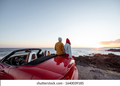 Couple enjoying beautiful views on the ocean, hugging together near the car on the rocky coast, wide view from the side with copy space on the sky