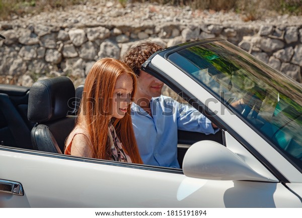 Couple enjoying a drive in a\
convertible in summer road. Friends going on holidays. Italian\
vacation serpentine roads, a redheaded woman and a beautiful young\
man