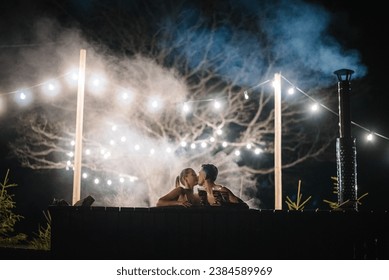 Couple enjoying hot tub at night outdoors. Winter holidays in mountains. Family relax in thermal spa open air. Man and woman swims in hot bath while resting. Glowing garlands and light bulbs. - Powered by Shutterstock