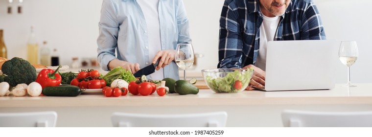 Couple Enjoying And Preparing Healthy Meal In Kitchen And Reading Recipes On Laptop. Happy Adult Woman Makes Salad Of Vegetables, Man Searches Class In Internet At Kitchen Interior, Empty Space