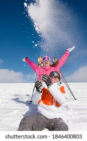 Couple enjoying while sitting down on a ski slope next to their sports gear as they throw snow in the air