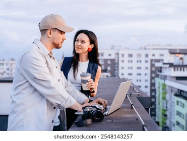 Couple enjoys working outdoors on rooftop with laptop and camera. Man and woman share ideas during sunset. Modern urban life teamwork, creativity. Collaborative atmosphere. - Powered by Shutterstock