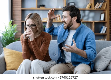 Couple experiencing relationship problems, a frustrated and enraged man yelling aggressively at his distressed and upset wife sitting on the couch, causing emotional distress, abuse, and sadness