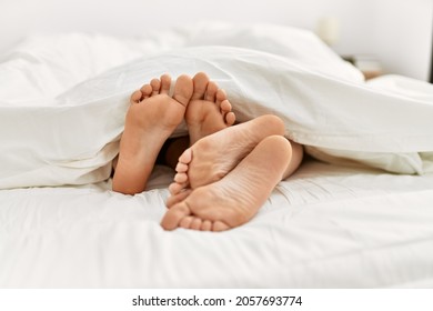 Couple Feet Under Sheets On The Bed At Home.