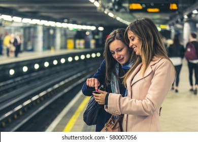 Couple of female friends looking at the smartphone while waiting for the train on the tube. Two young women checking on the phone before boarding. Tone added