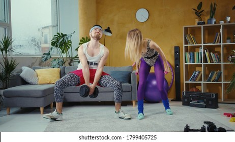 Couple Of Fitness Jerks Going In For Sports At Home Lifting Up Weights. Joyful Retro Style Woman Making Fun Of Struggling Bearded Man Exersicing With Heavy Dumbbells.