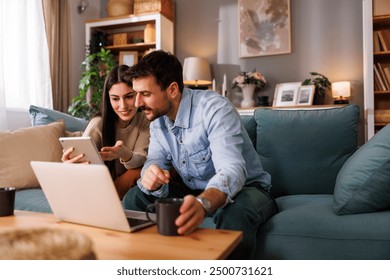 Couple of freelance IT specialists sitting on living room sofa using digital technology devices while working remotely from home, woman using tablet computer while man working on laptop computer - Powered by Shutterstock