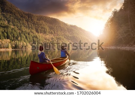 Couple friends canoeing on a wooden canoe during a colorful sunny sunset. Cloudy Sky Composite. Taken in Harrison River, East of Vancouver, British Columbia, Canada.