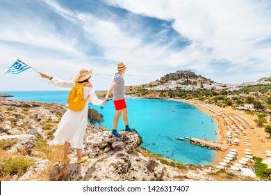 A couple of friends in love with the Greek flag admire the grandiose view of the sea Bay and the beach near the ancient city of Lindos. Honeymoon and travel - Powered by Shutterstock