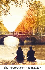 Couple friends or lovers relaxing on embankment of canal and looks at the river and bridges of Amsterdam. Photo toning at retro color. Spring or autumn time in the city. Travel Netherlands, Europe.