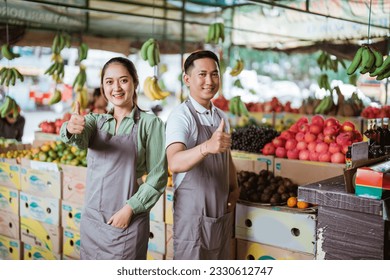 a couple of fruit seller with apron standing together with thumbs up with the fruits at the background - Powered by Shutterstock