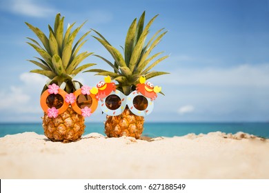 Couple Of Funny Attractive Pineapples In Children's Cheerful Sunglasses On Sand Against Turquoise Sea. Tropical Summer Vacation Concept. Happy Sunny Day On The Beach Of Tropical Island. 