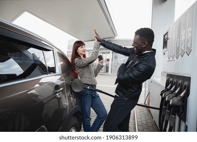 Couple At Gas Station. Happy Joyful Multiracial Couple Of Friends Or Colleagues, Having Fun While Refueling Luxury Car At Petrol Station, Giving High Five Each Other And Smiling