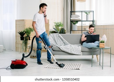 Couple Of Gay Cleaning Living Room At Home, Young Gay Man Holding The Laptop, Sitting Sofa. Young Gay With Apron Using Vacuum Holiday Together