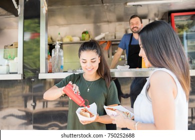 Couple of girls standing in front of a food truck and eating hot dogs with ketchup