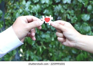 Couple hands holding two pieces of puzzle forming red heart on green leaves blur background, heart on pieces of jigsaw on lover hand, romantic lover celebrate on Valentine day, love unity of  couple