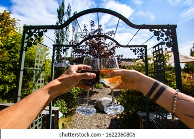 Couple Hands Toasting And Cheering Red And White Wine Glasses In The Front Of A Winery Garden Gate, Tasting Restaurant Patio In The Background