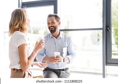 Couple of happy colleagues talking while standing at the office window with cups of coffee