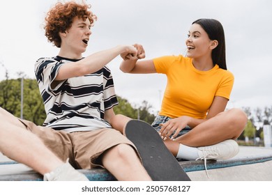 Couple of happy friends teenagers boy girl classmates high school pupils students give fist bump celebration have fun looking to each others yelling sitting in skate ramp park riding on skateboard - Powered by Shutterstock