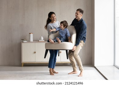 Couple of happy funny parents holding cheerful little son sat in armchair. Family moving into new house, carrying furniture, feeling joy. Mom and dad playing active games with kid. Relocation concept