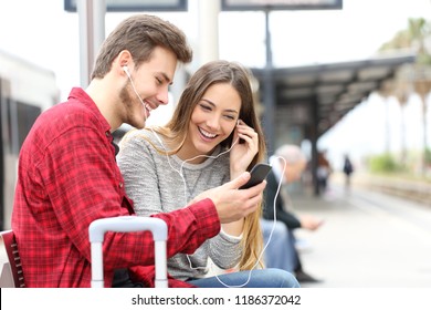 Couple of happy travelers waiting in a train station sharing music with a smart phone and earphones - Powered by Shutterstock