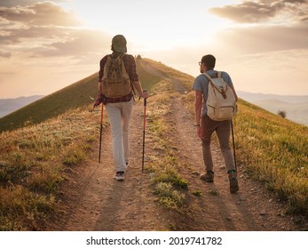 A Couple Of Happy Young Hikers Backpacking On A Beautiful Rocky Trail On A Warm, Sunset Evening. The Concept Of Family Travel And Adventure. View From Behind. Travel Lifestyle.