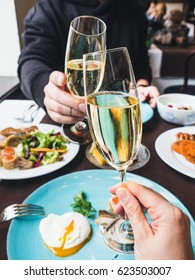 Couple having a brunch with sparkling wine in a fancy hotel or restaurant. Glasses of sparkling wine in hands, cheers!  Benedict  egg and other food on a background