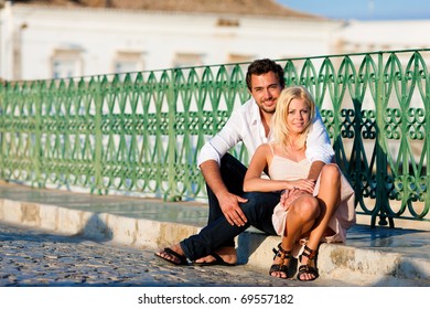 Couple Having A City Break In Summer Sitting On A Bridge Over A River In The Evening Light