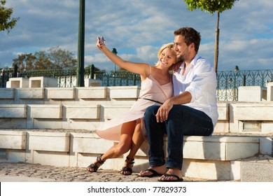 Couple Having A City Break In Summer Sitting On A Brick Wall In The Sunlight, Making Pictures