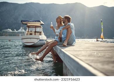 Couple having a drink and laughing by seaside sitting on wooden jetty by water. Holiday, lifestyle, togetherness concept. - Powered by Shutterstock