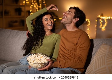 Couple Having Fun Eating Popcorn, Girlfriend Feeding Boyfriend Flirting During Date Sitting On Couch At Home. Young Spouses Enjoying Weekend Together. Romantic Relationship, Love And Friendship - Powered by Shutterstock