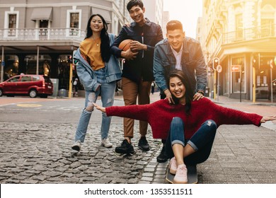 Couple having fun on skateboard with friends walking by outdoors. Man pushing woman on skateboard with their friends walking by in the city. - Shutterstock ID 1353511511