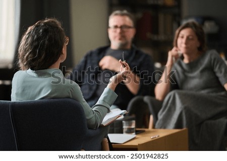 Couple having therapy session with female therapist gesturing while holding pen, room with bookshelf and therapy materials, focus on conversation, atmosphere calm and professional