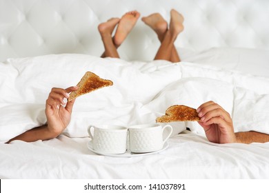 Couple Hiding Under Duvet Enjoying Breakfast In Bed