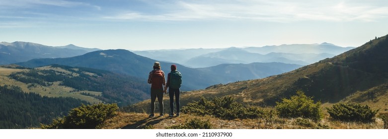 Couple of hikers with backpacks enjoying valley landscape view from top of a mountain. Young adult tourists, man and woman standing on the pass and holding by hands. Panoramic view of mountain hills - Powered by Shutterstock