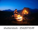 Couple hikers enjoys cozy moment by campfire, sipping hot drinks near glowing tent. Twilight sky and mountain backdrop create serene and romantic atmosphere in picturesque wilderness setting.