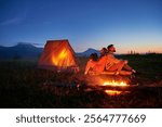 Couple hikers enjoys cozy moment by campfire, sipping hot drinks near glowing tent. Twilight sky and mountain backdrop create serene and romantic atmosphere in picturesque wilderness setting.