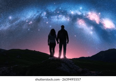 Couple of hikers holding hands standing on a rocky outcrop in mountains, night pink starry sky with glowing Milky Way. Colorful landscape with silhouettes of man and woman, bright stars and milky way - Powered by Shutterstock - Get 15% off with code: PIKWIZARD15