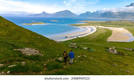 A couple hikes a grassy path in Norway, enjoying a panoramic view of a pristine, crescent-shaped beach and the rugged mountains beyond. Lofoten Norway - Powered by Shutterstock