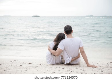 Couple holding each other on beach. Young happy interracial couple on beach holding each other sitting down. Asian woman, Caucasian man. Young mixed race romance concept.