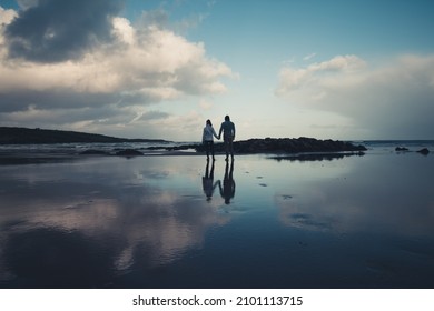 A Couple Holding Hands On A Beach During Winter. Sharing Their Love As They Look Out Onto The Ocean. A Strong Reflection Of The Couple In The Soaked Beach Lies Behind Them. 