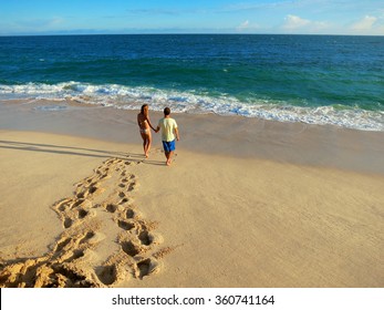 Couple Holding Hands Walking On Beach To The Ocean. Man And Woman In Love. Footprints In The Sand.