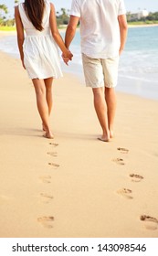 Couple holding hands walking romantic on beach on vacation travel holidays leaving footprints in the sand. Closeup of feet and golden sand for copy space. Young couple wearing white shorts.