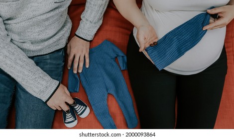 

Couple Holding Newborn Baby's Things While Sitting On Sofa, Top View Close-up.