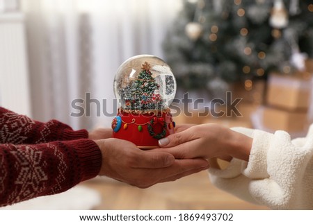Couple holding snow globe with Christmas tree at home, closeup