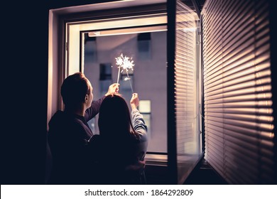 Couple Holding Sparklers Out Of The Window At Night. New Year's Eve Celebration, Anniversary, Party Or Date At Home. Spontaneous Candid Fun With Light Firework Stick. Happy And Playful Romantic Moment