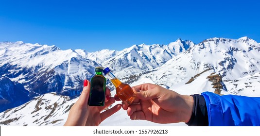 A Couple Holding Two Different Bottles With Liquor And High Snowy Mountains In The Back. Woman's Nails Are Painted Red. Apres Ski In Austrian Alps, Heilgenblut.