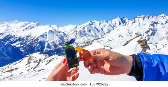 A Couple Holding Two Different Bottles With Liquor And High Snowy Mountains In The Back. Woman's Nails Are Painted Red. Apres Ski In Austrian Alps, Heilgenblut.