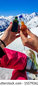 A Couple Holding Two Different Bottles With Liquor And High Snowy Mountains In The Back. Woman's Nails Are Painted Red. Apres Ski In Austrian Alps, Heilgenblut.