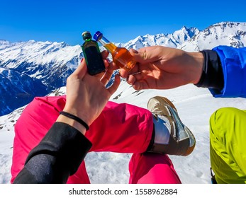 A Couple Holding Two Different Bottles With Liquor And High Snowy Mountains In The Back. Woman's Nails Are Painted Red. Apres Ski In Austrian Alps, Heilgenblut.